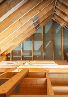 A carpenter carefully measures and adjusts wooden beams in an attic, illuminated by sunlight filtering through the roof. The workspace reflects a renovation effort.