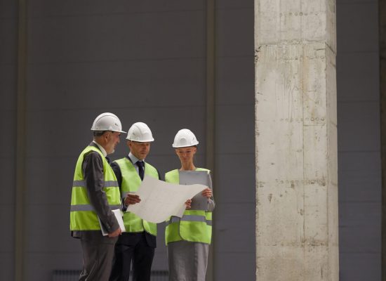 Vertical full length portrait of business people wearing hardhats and holding plans while standing at construction site indoors, copy space